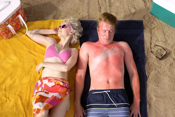 Man looking at his sunburned chest, with the imprint of his wife's arm beside him as they lie on the beach